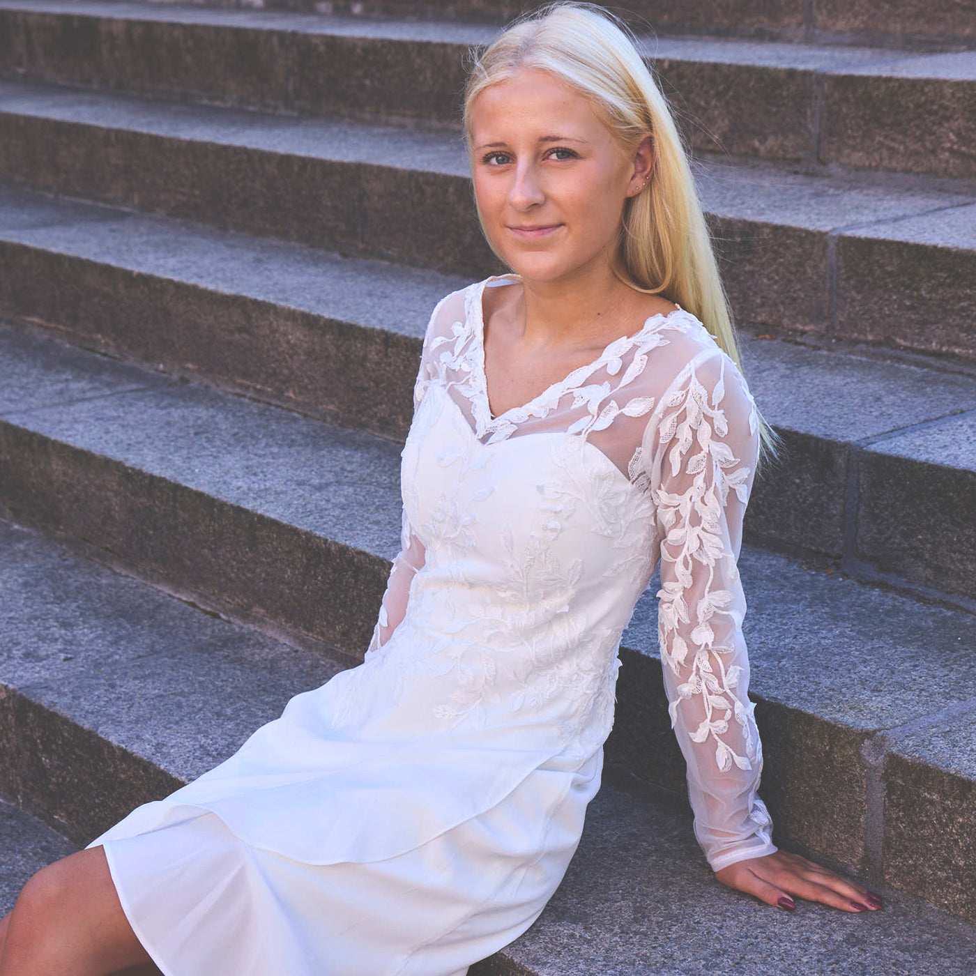 Pige sidder tilbagelænet på trappe i hvid konfirmationskjole. Overdelen er i blød tyl med fin V-neck foran. Smukke blonde applikationer på overdelen og ned langs ærmerne. Kjolen har en asymmetrisk flæsedetalje på chiffon-underdelen.